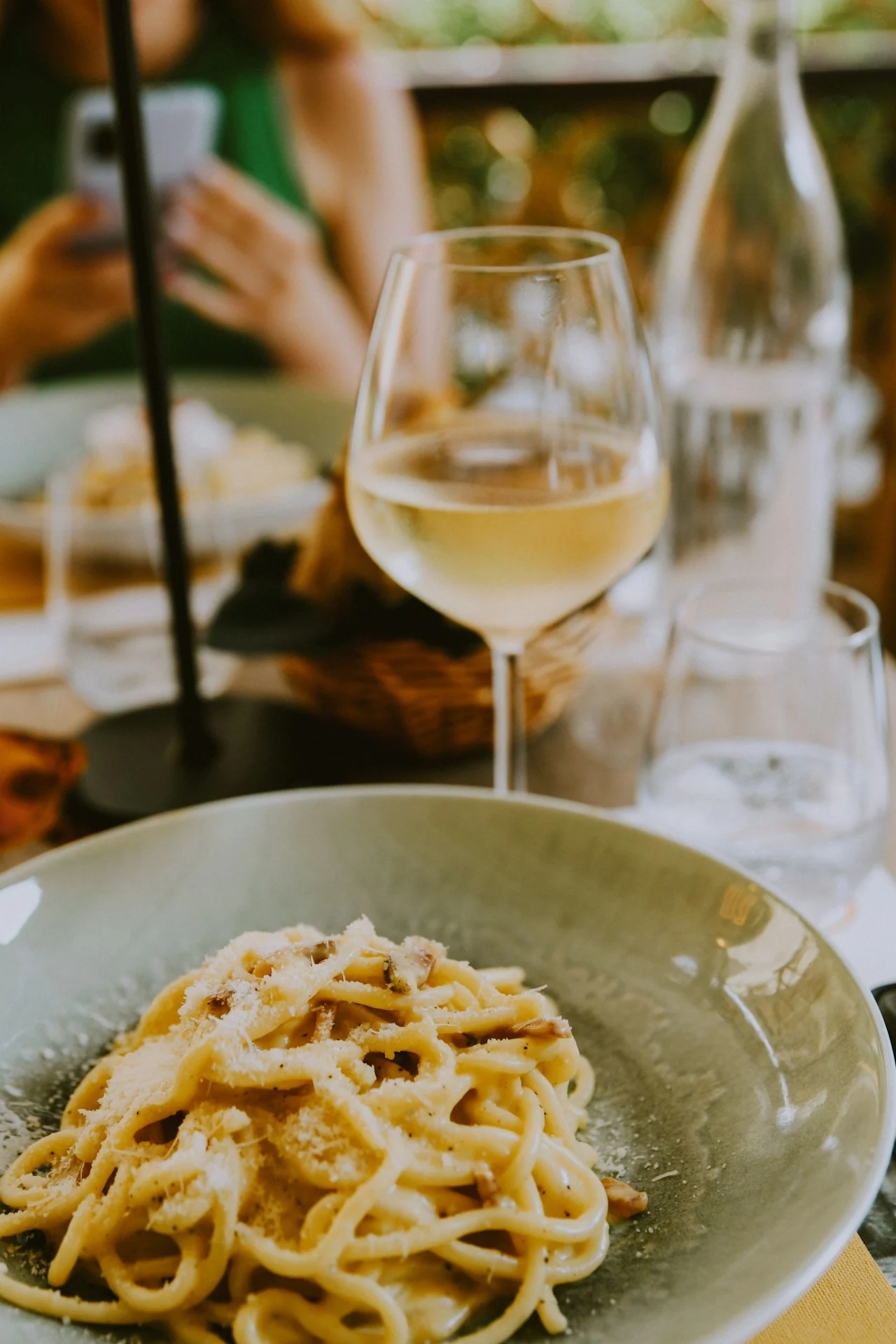 A plate of creamy ribbon pasta topped with grated cheese beside a glass of white wine at an Italian table
