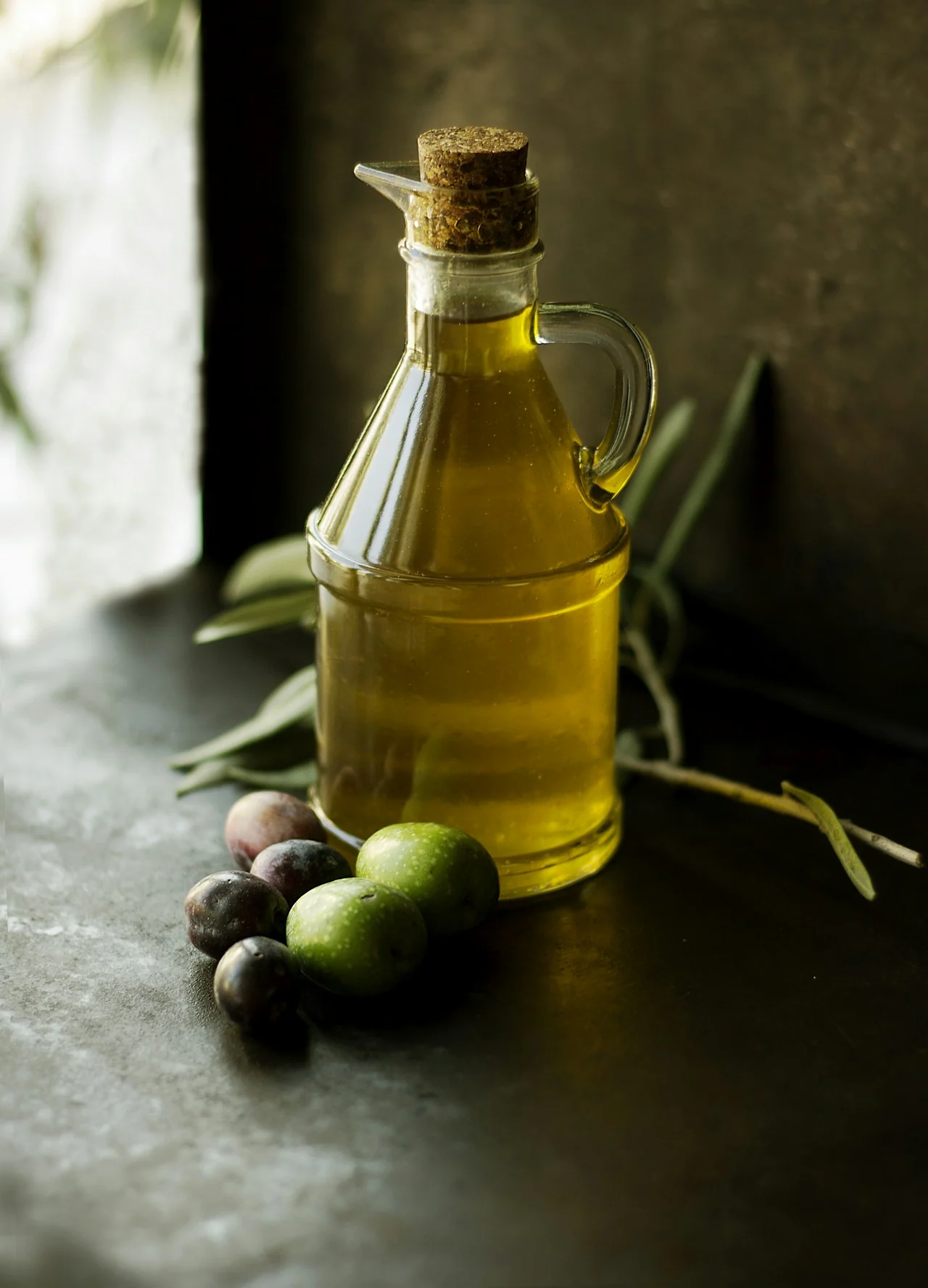 A corked glass cruet of golden olive oil beside green and purple olives in soft window light
