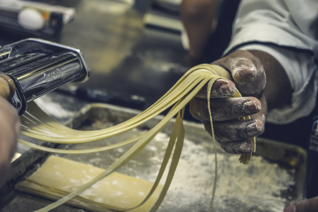 Flour-dusted hands lifting fresh ribbons of pasta from a pasta machine