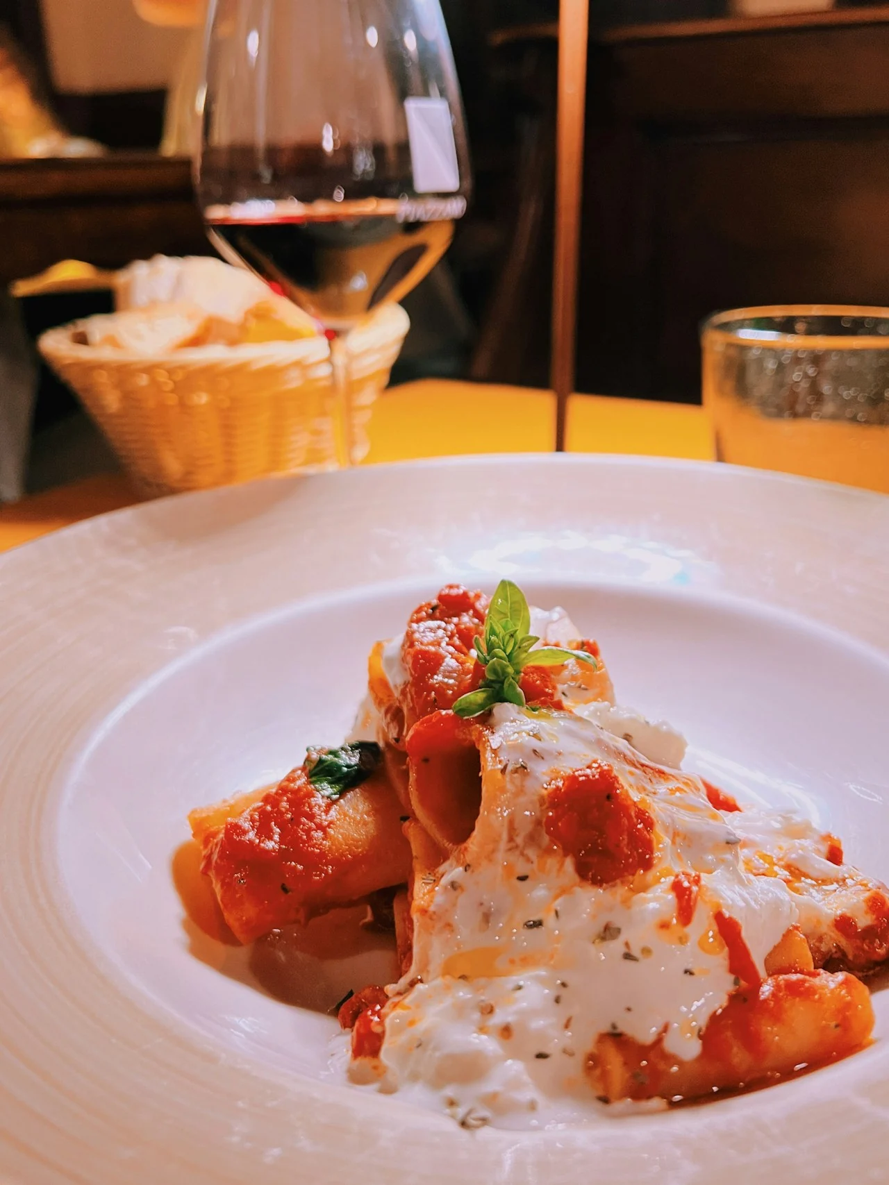 Große Röhrennudeln mit Tomatensauce, geschmolzenem Käse und Basilikum neben einem Weinglas und einem Brotkorb