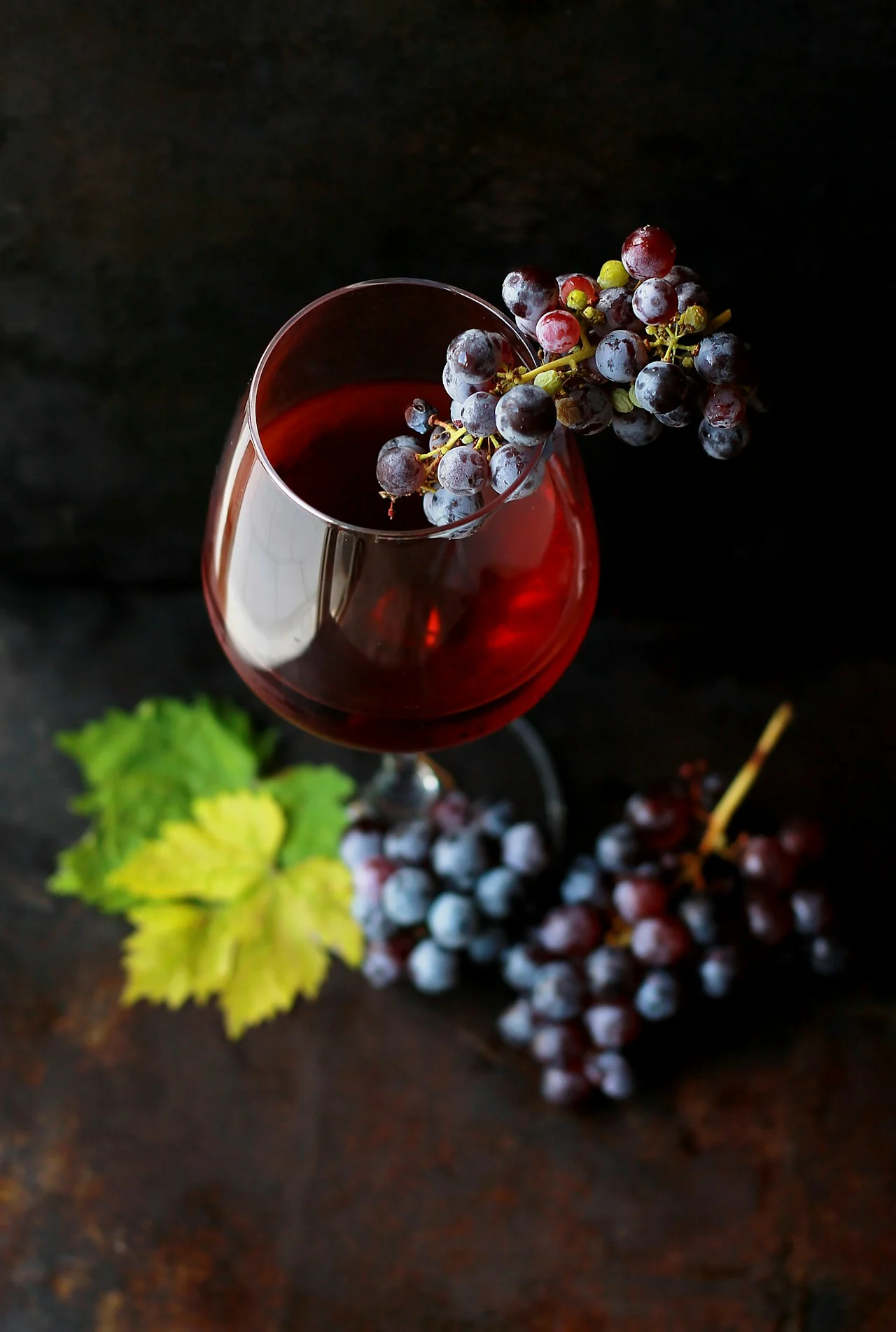 A glass of red wine with dark grapes and a green vine leaf on a dark surface