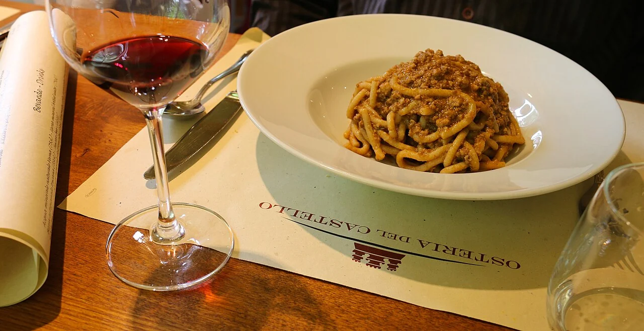 Tuscan pici pasta with Chianti-style ragù beside a glass of red wine on a rustic restaurant table