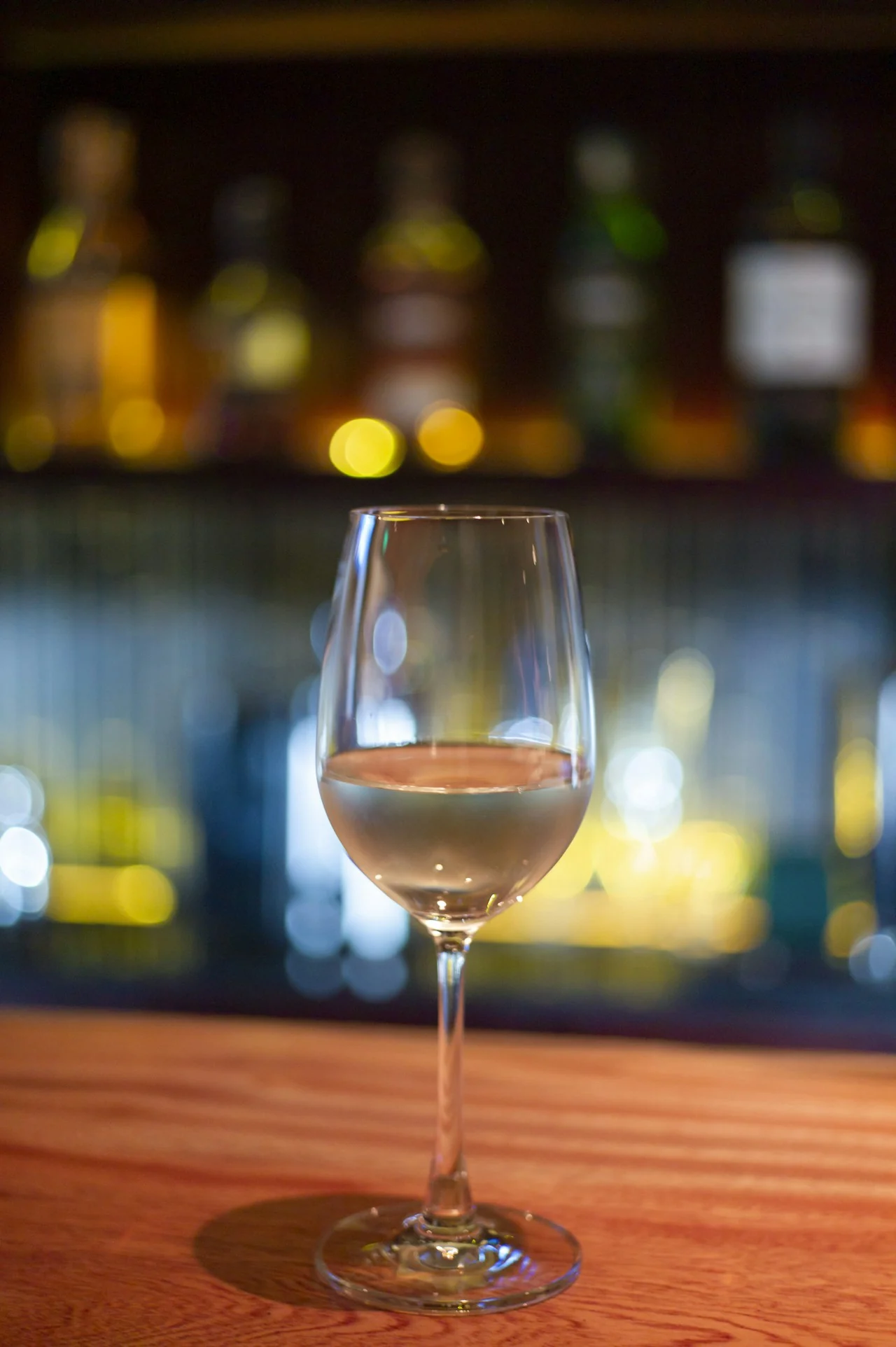 A glass of pale wine on a wooden counter against softly blurred coloured lights